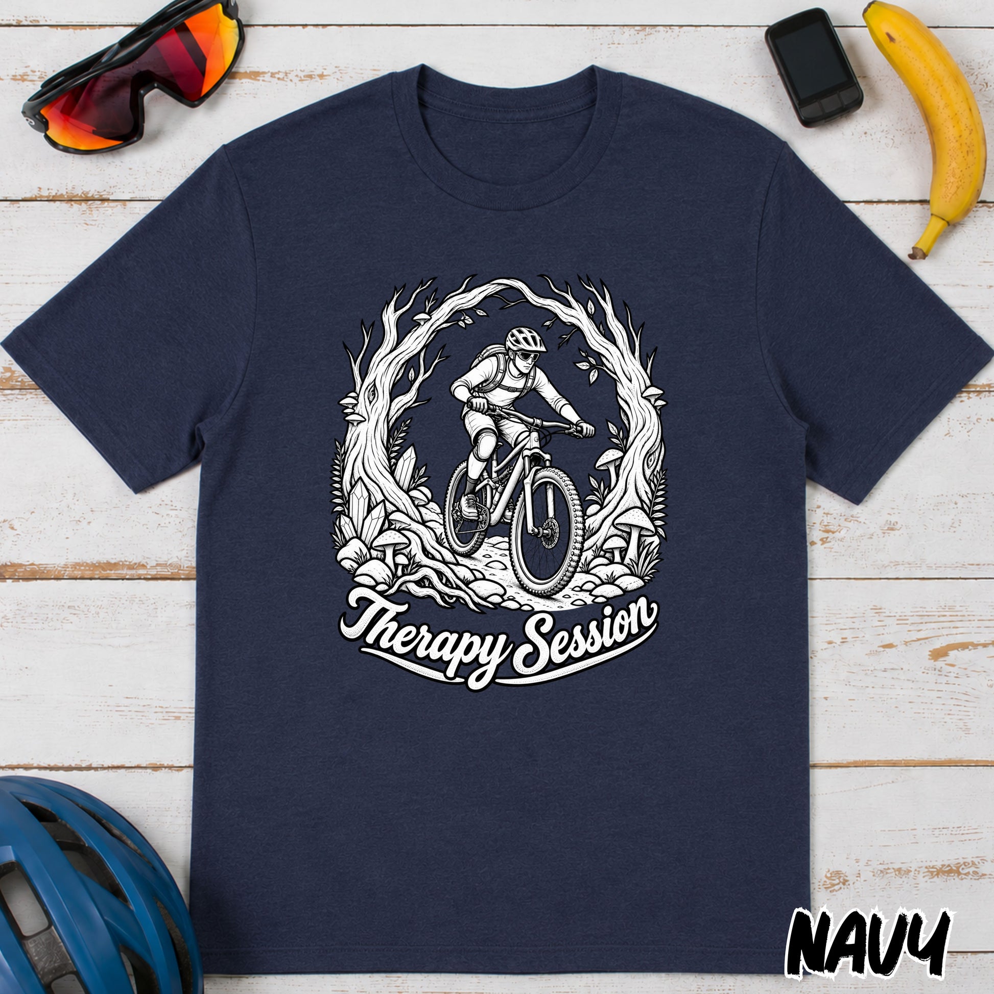 A navy blue short-sleeve t-shirt with a white graphic of a mountain biker inside a circular wreath and the text 'Therapy Session' below it, laid flat on a white wood background with lifestyle props including sunglasses, a phone, a banana, a helmet, and the word 'NAVY' in the corner.
